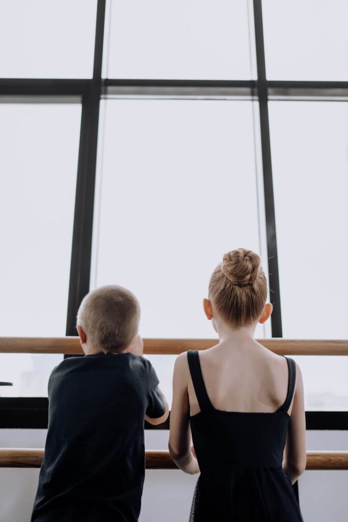 Two children, a boy and a girl in ballet attire, gaze out a large window indoors.